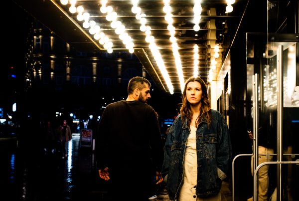 A man and a woman stood outside a bar at night. The woman is looking straight ahead while the man has his head turned looking at her